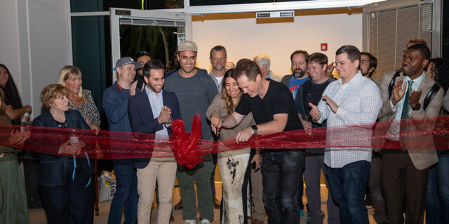 A group of people excitedly cut a sheer red ribbon.