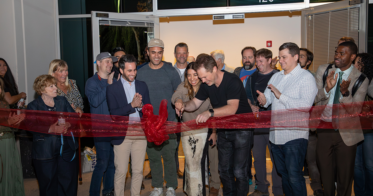 A group of people excitedly cut a sheer red ribbon.