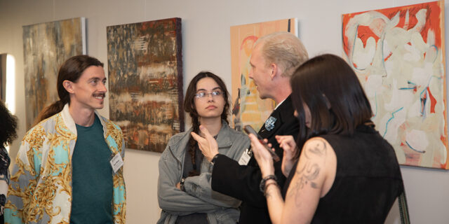 Two men and two women stand in front of large art pieces on the wall behind them, speaking to each other.