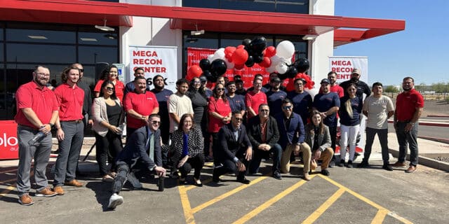 A group of more than 30 people pose in front of a balloon arch and warehouse facility.