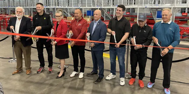 A line of people stand behind a red ribbon with oversized scissors in the PUMA distribution/warehouse facility in Glendale, Ariz.