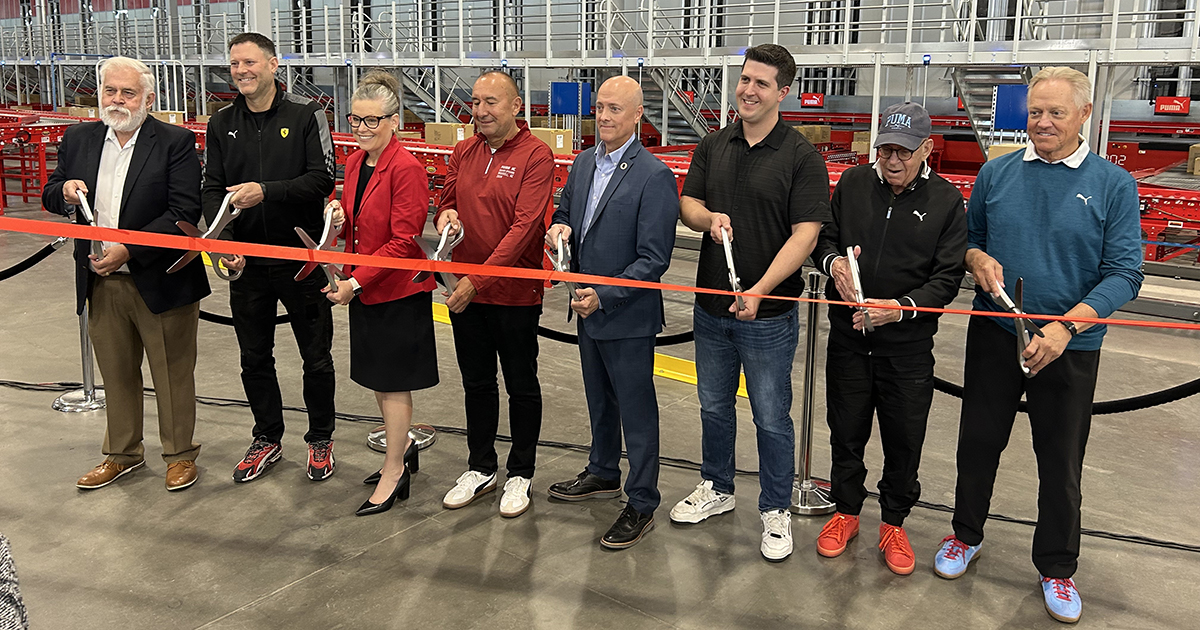 A line of people stand behind a red ribbon with oversized scissors in the PUMA distribution/warehouse facility in Glendale, Ariz.