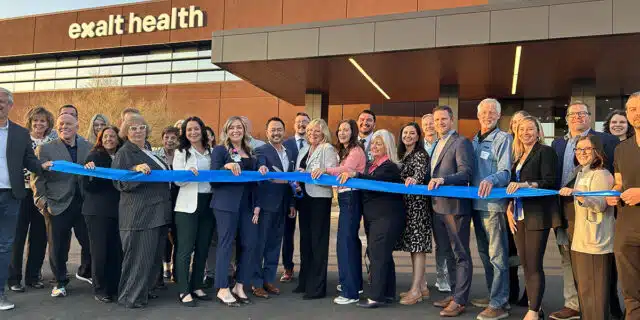 A group of people stand with a blue ribbon in front of the Exalt Health building.