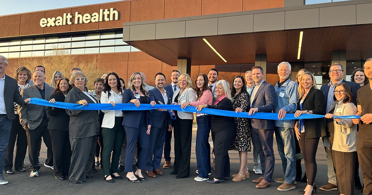 A group of people stand with a blue ribbon in front of the Exalt Health building.