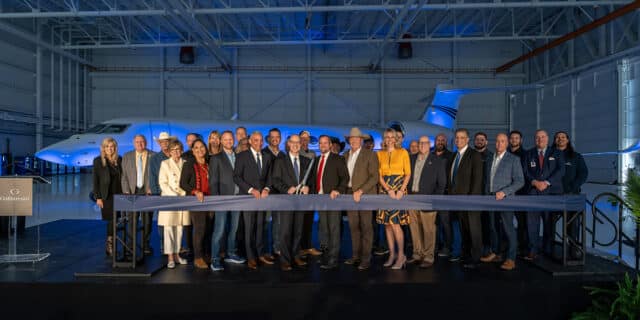 A gaggle of people stand behind a thick dark ribbon, with the person in the middle holding giant scissors. Behind them is an airplane.