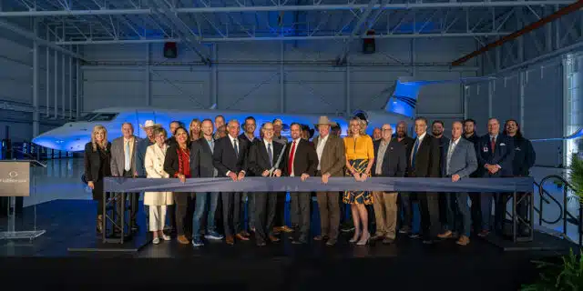 A gaggle of people stand behind a thick dark ribbon, with the person in the middle holding giant scissors. Behind them is an airplane.