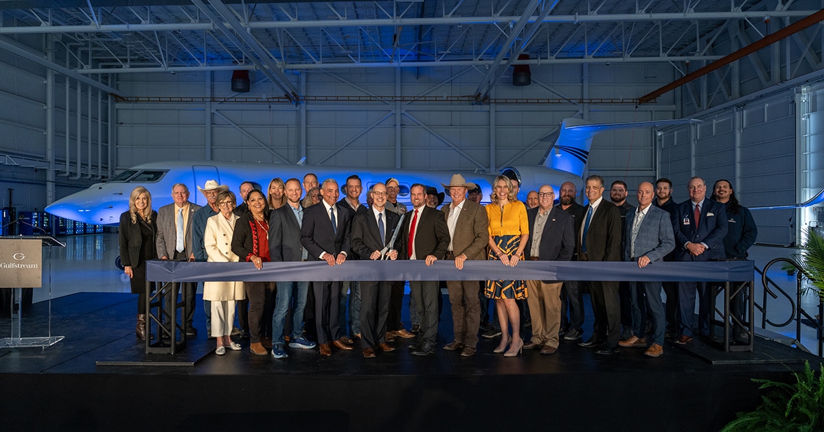 A gaggle of people stand behind a thick dark ribbon, with the person in the middle holding giant scissors. Behind them is an airplane.